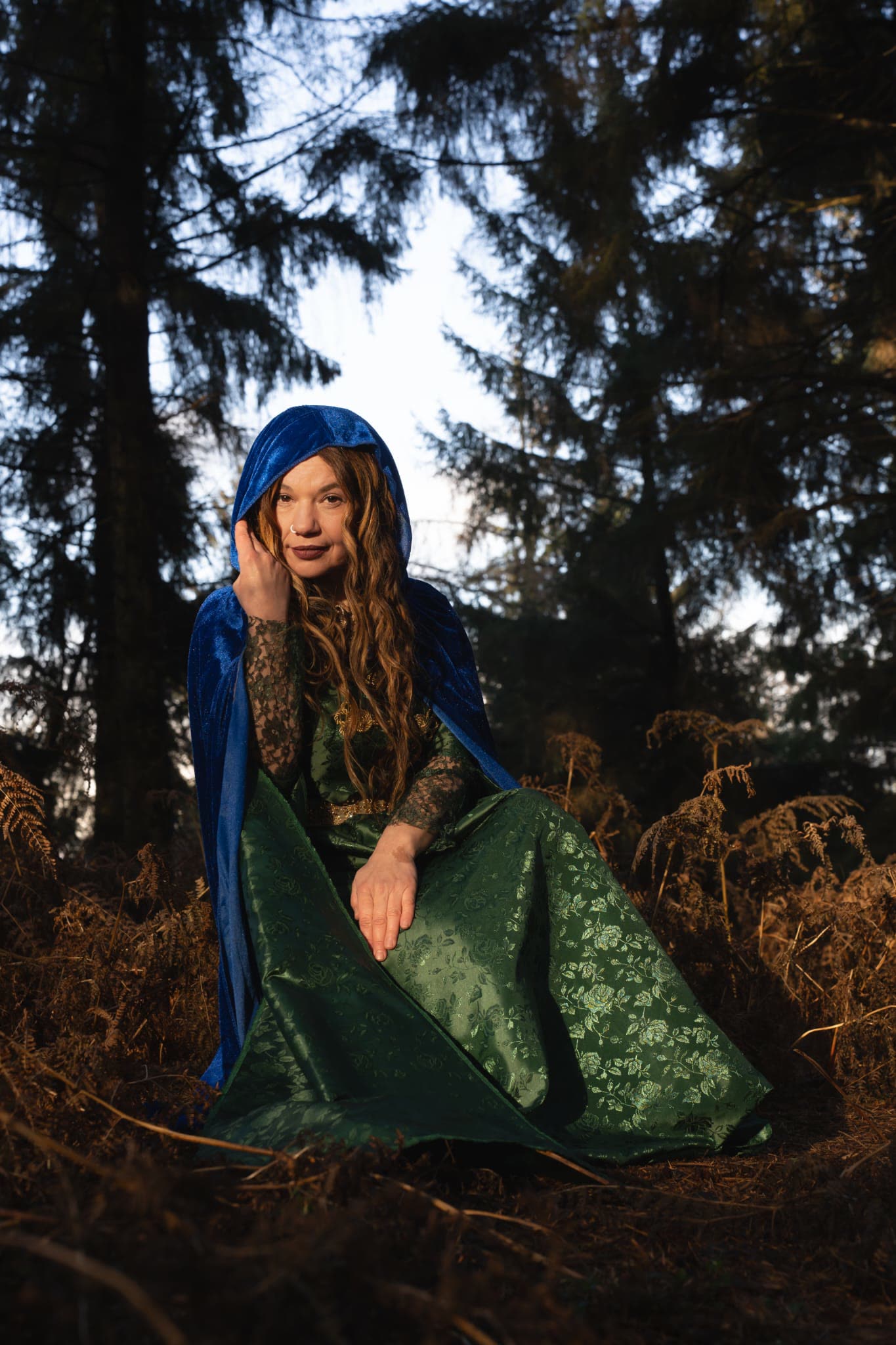 Aynaz wrapped in a blue velvet cloak beneath the pines